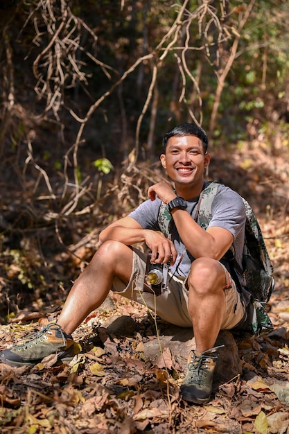 Handsome Asian man with backpack and film camera sits on a rock in the forest resting after hiking