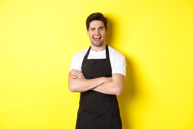 Handsome barista in black apron winking at you, wearing black apron uniform, standing over yellow background.