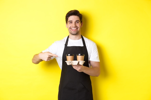 Handsome barista holding two cups of takeaway coffee, pointing finger at drinks and smiling, standing in black apron against yellow background