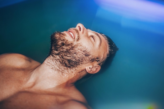 Handsome beard man floating in tank filled with dense salt water used in meditation, therapy, and alternative medicine.