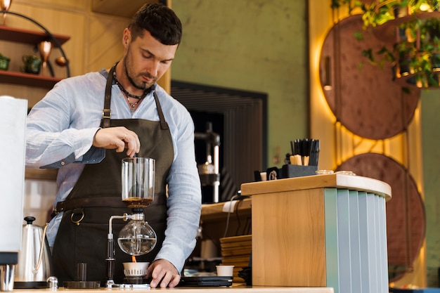 Handsome bearded barista mix freshly ground coffee in syphon device for coffee brewing in cafe