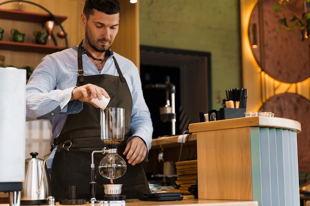 Handsome bearded barista pours freshly ground coffee to syphon device for coffee brewing in cafe