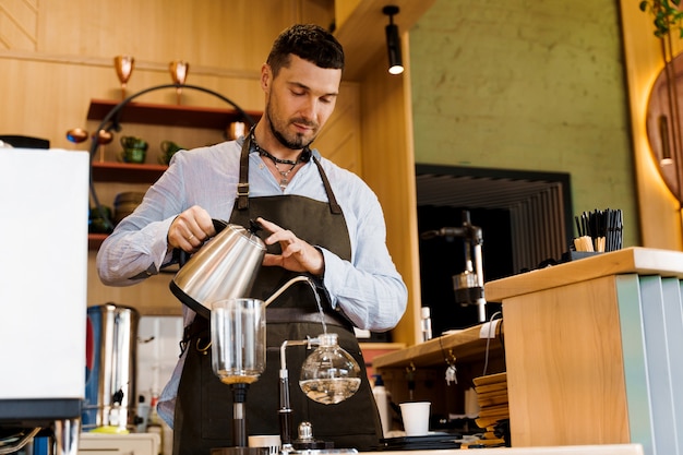 Handsome bearded barista pours hot boiling water from kattle to syphon device for coffee brewing in cafe