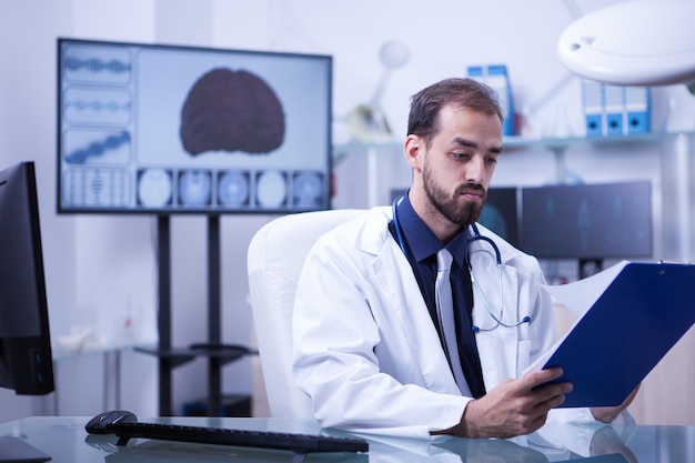 Handsome bearded doctor checking the list of patients to visit for the day. Monitor with 3d brain in the background.