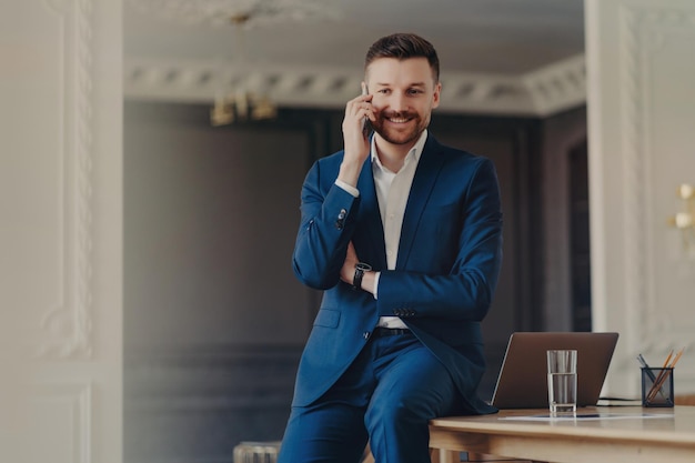 Handsome bearded man in blue suit satisfied with good news talking on the phone posing at desk with modern laptop Mobile conversation