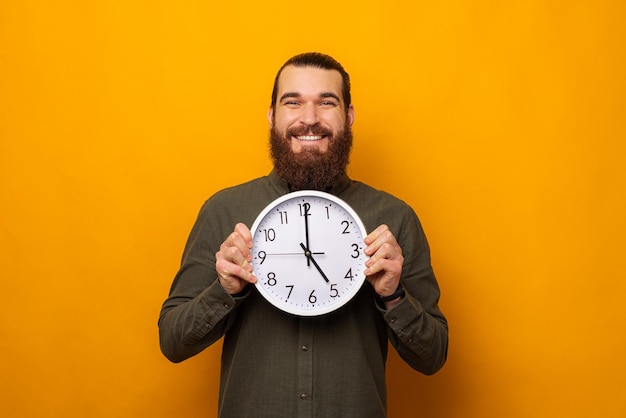 Handsome bearded man is holding a round white wall clock while smiling