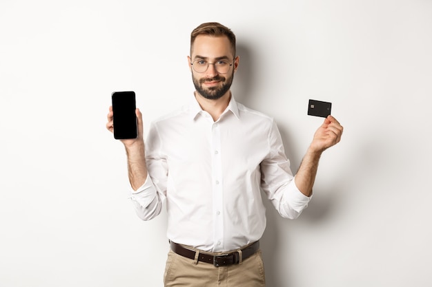 Handsome bearded man showing mobile phone and credit card, shopping online, standing over white background.