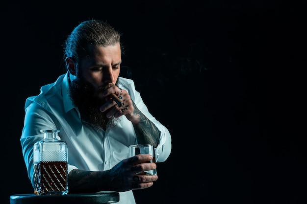 Handsome bearded man smoke a cigar holds glass of whiskey dressed in white shirt studio shot