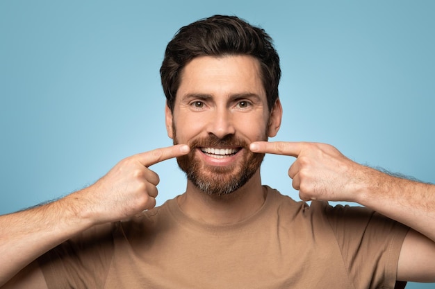 Handsome bearded man in tshirt posing over blue background smiling and pointing with fingers at perfect teeth