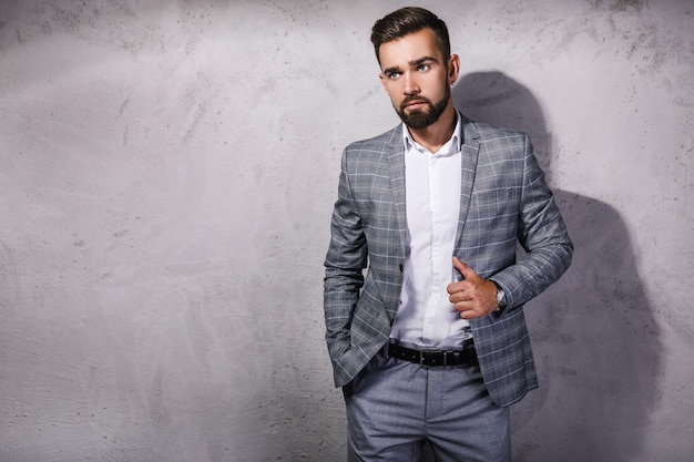 Handsome bearded man wearing gray suit is posing against concrete wall