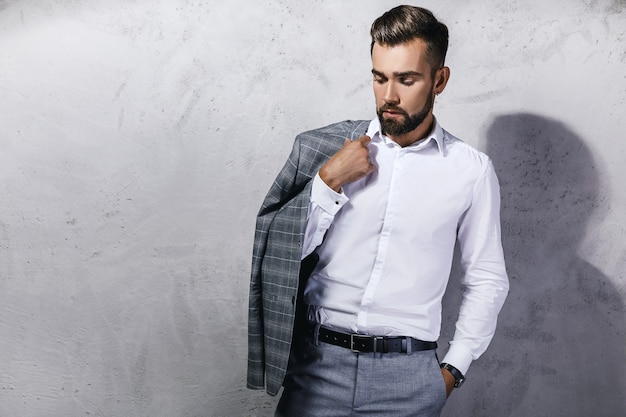 Handsome bearded man wearing gray suit is posing against concrete wall