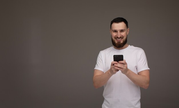 Handsome bearded model is posing with phone in hands over grey background.