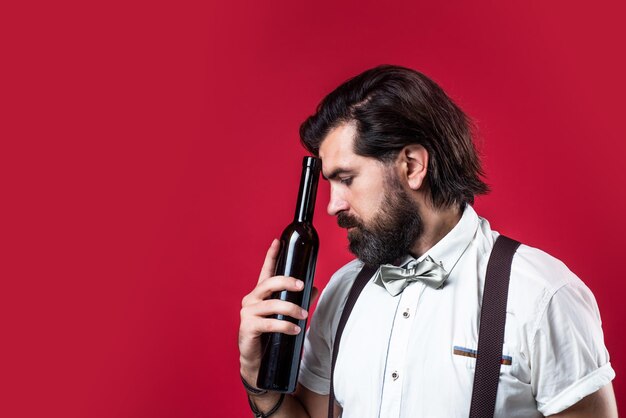 Handsome bearded rich man with stylish hair mustache and beard in suspenders holding bottle of wine sommelier