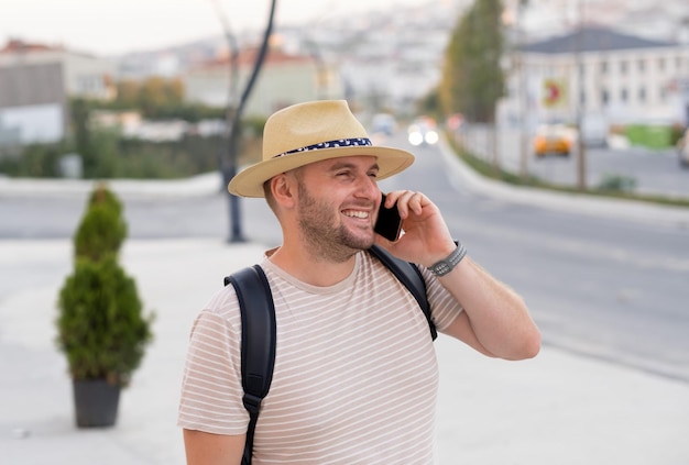 Handsome bearded smiling mantourist wearing sunhat backpack having call using smartphone in city outdoorsHappy caucasian guy speaking by phone travelling