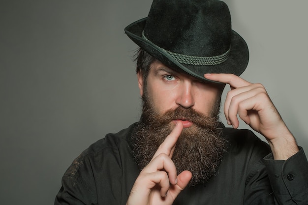 Handsome bearded young man hipster or gentleman with long beard and mustache in stylish black retro hat on serious face in studio on grey background portrait