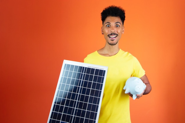 Handsome Black Man Holding a Piggy Bank and Solar Panel Isolated on Orange Man in a yellow shirt isolated