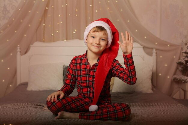 a handsome blond boy in a Santa Claus hat in Christmas pajamas is sitting on the bed