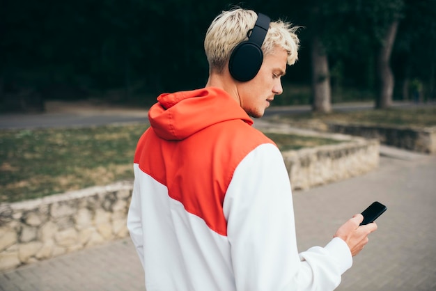 Handsome blond hipster guy listening to audiobook using his headphones and mobile phone messaging via social networks using wireless Internet connection during walk in the park Street fashion