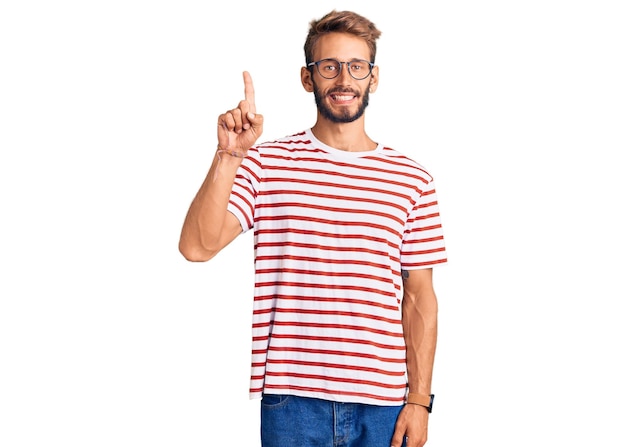 Handsome blond man with beard wearing casual clothes and glasses showing and pointing up with finger number one while smiling confident and happy