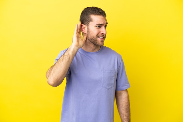 Handsome blonde man isolated listening to something by putting hand the ear