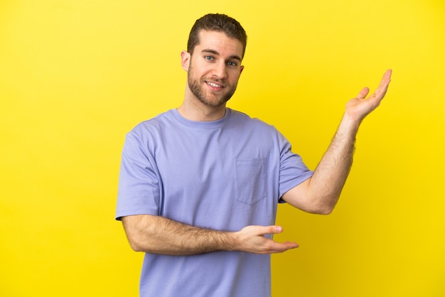 Handsome blonde man over isolated yellow background extending hands to the side for inviting to come