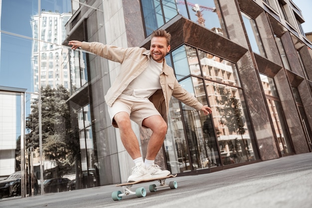 Handsome blonde young man doing tricks on skateboard trains on longboard outdoors at street with urban background. Excited laughing sporty skater practices outside. Extreme sport lifestyle concept.