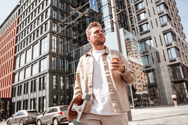 Handsome blonde young man skateboarder holding longboard and cup of coffee takeaway looks seriously focused while crossing the road on urban building background. Wears casual denim outfit.