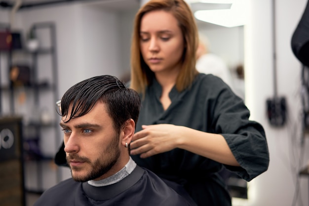 Handsome blue eyed man sitting in barber shop. Hairstylist Hairdresser Woman cutting his hair. Female barber.