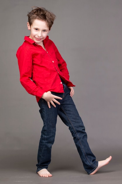 Handsome boy in a red shirt and jeans on a gray wall. A child in the modeling business is posing.