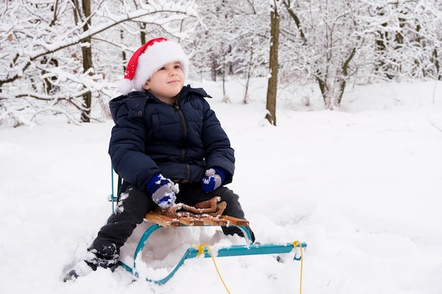 A handsome boy in a Santa Claus hat sits on a sled