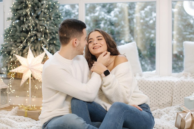 Handsome boy with his beautiful girlfriend hug each other at home at christmas eve fit tree and snow