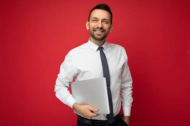 Handsome brunet man holding laptop computer looking up in white shirt and tie on isolated red wall.