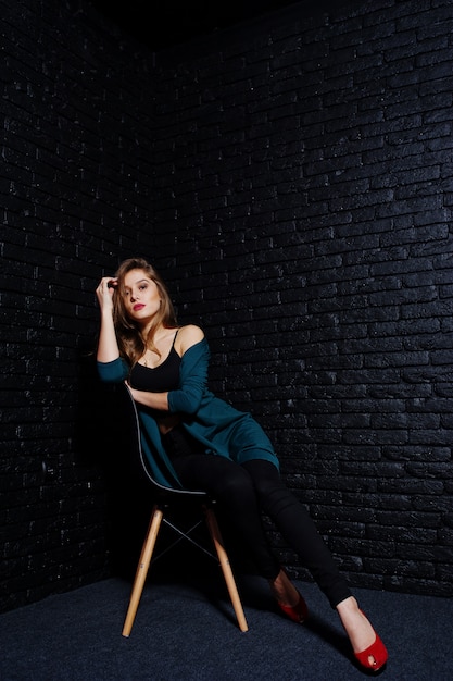 Photo handsome brunette girl wear on black and green jacket in red high heels, sitting and posing on chair at studio against dark brick wall.