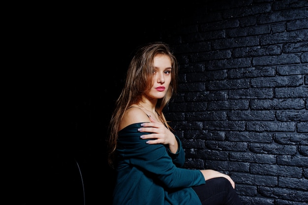 Handsome brunette girl wear on black and green jacket, sitting and posing on chair at studio against dark brick wall.