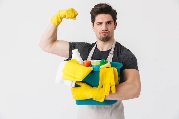 Handsome brunette houseman wearing apron standing isolated over white , holding bucket full of detergents