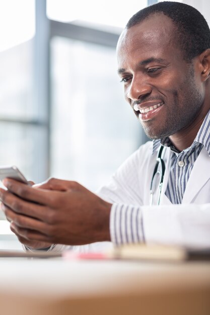 Handsome brunette male keeping smile on face while leaning arms on table and looking at telephone