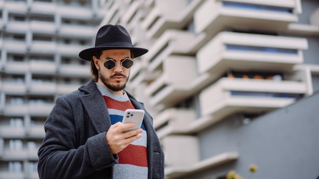 Handsome brunette man in hat and sunglasses using smartphone while standing outdoors in the city