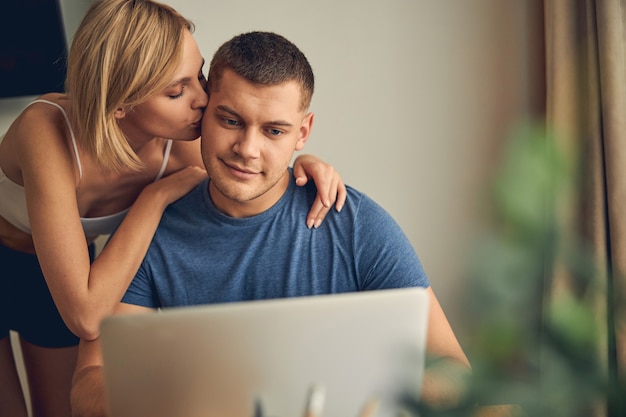 Handsome brunette man working online on computer while blond slim woman hugging and kissing him