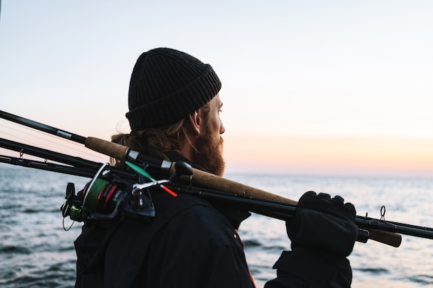 Handsome brutal bearded fisherman wearing coat standing with a fishing rod at the seashore