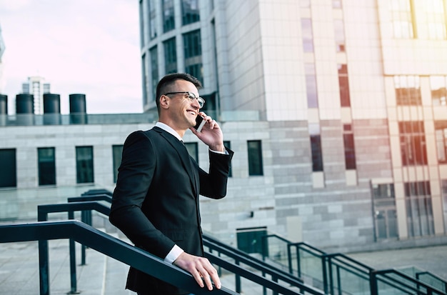 Handsome businessman in full black suit talks on his smart phone at the cityscape background