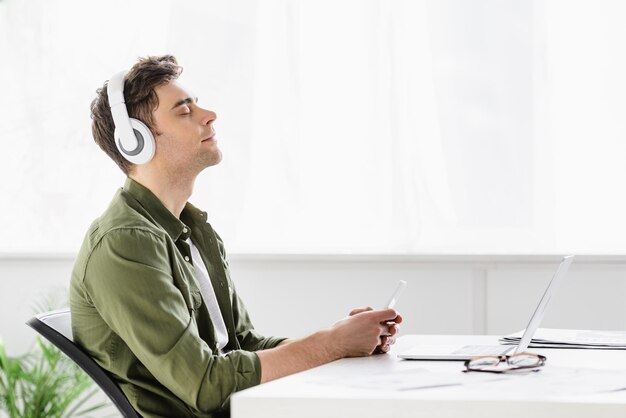 Photo handsome businessman in headphones sitting near table with laptop holding smartphone and listening