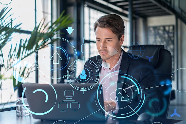 Handsome businessman in suit at workplace working with laptop to optimize development by implying new technologies in business process Hi tech hologram over office background