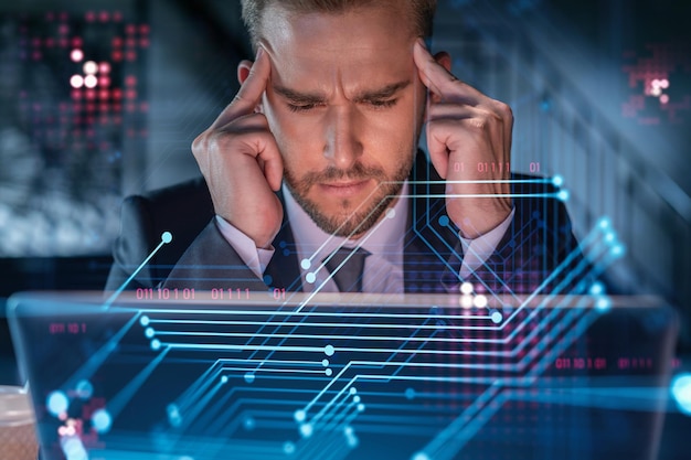 Handsome businessman in suit at workplace working with laptop to optimize development by implying new technologies in business process Hi tech hologram over office background