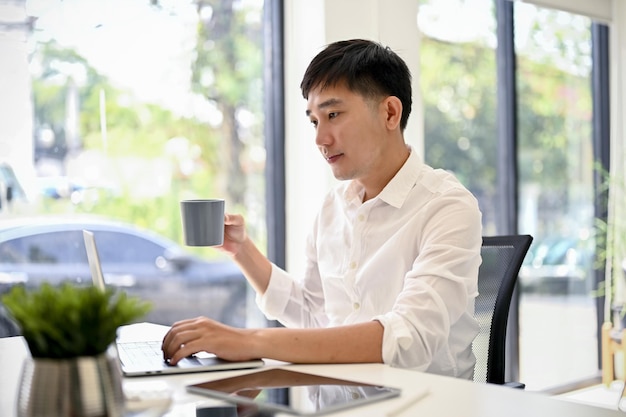 Handsome businessman using his laptop and sipping coffee at his desk in a modern office