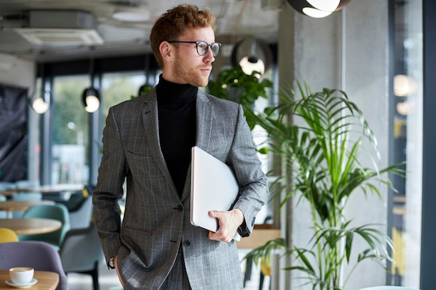 Handsome businessman wearing stylish suit and eyeglasses holding laptop looking away, copy space