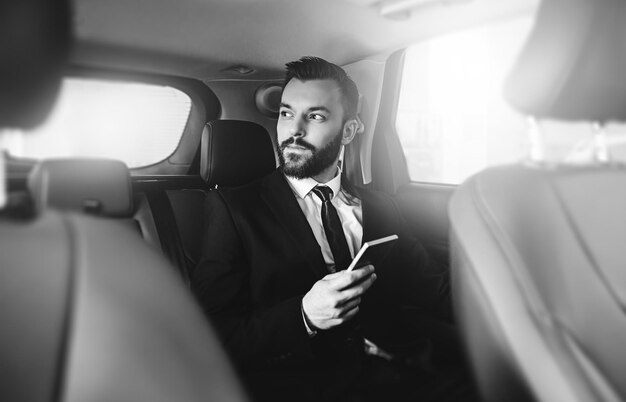 Handsome businessman with phone on hand sitting on the backseat of the car guy looking away