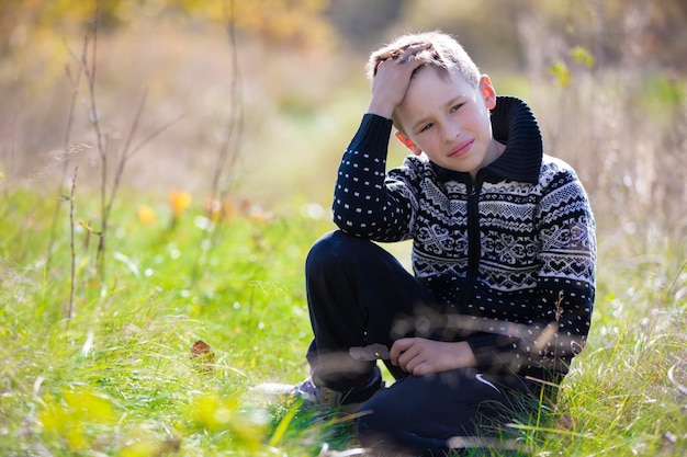 A handsome calm boy in a knitted sweater sits on the grass on the field