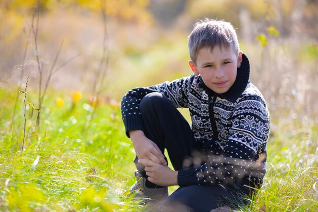 A handsome calm boy in a knitted sweater sits on the grass on the field