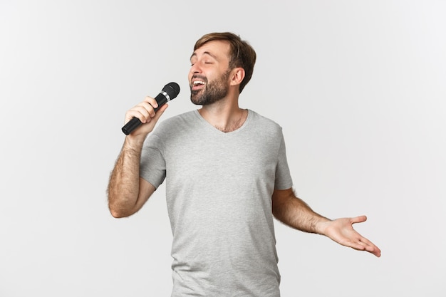 Handsome carefree guy singing song in karaoke, holding microphone, standing over white background