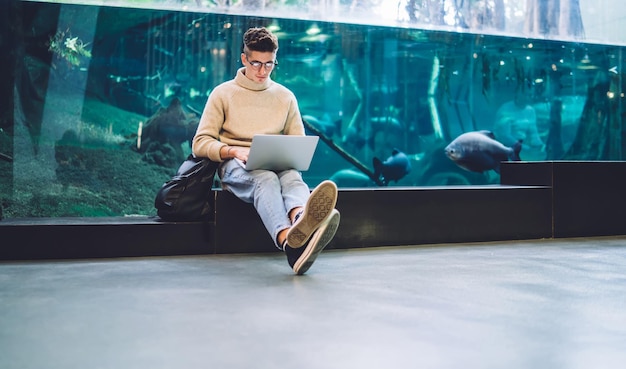 Handsome casual man in eyeglasses and casual outfit sitting with legs crossed typing on laptop